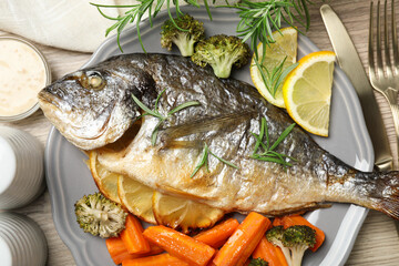 Cooked fish served with rosemary, lemon and vegetables on wooden table, flat lay