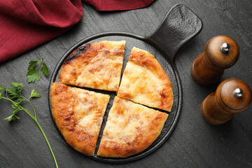 Cut delicious Megrelian khachapuri, spices and parsley on black table, flat lay