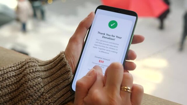 A moment of digital philanthropy captured in real-time. Hands hold a smartphone displaying a confirmed donation notification in a public setting, representing modern charitable giving through mobile