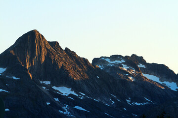 Mountain landscape with rocky, snowy peaks at sunset, lit by golden sunlight Fjords style