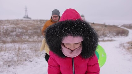 A beautiful girl in a pink jacket with a black hood walks towards the camera. Behind her in the bokeh are her dad and mom. Winter fun in a snowy nature.
