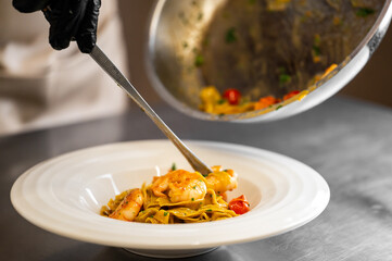 Chef plating shrimp pasta with cherry tomatoes and herbs in a professional kitchen. Close-up of gloved hand, spoon, and white plate on stainless steel surface.