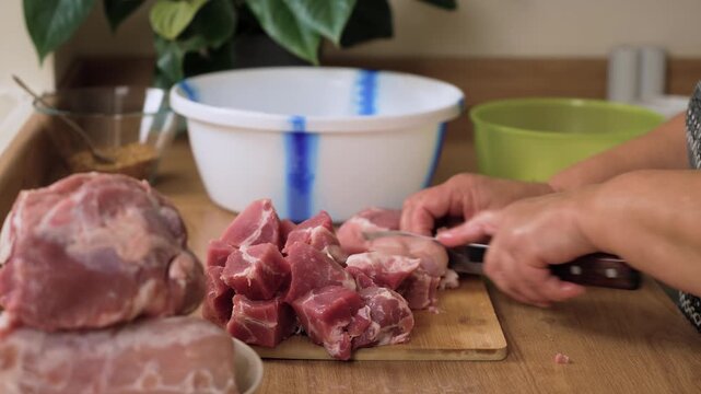 Slicing pork into medium cubes on wooden board. Unrecognizable person cutting meat for future marination. Fresh pieces are being prepared for onion spice mix. Cooking dish traditional shashlik skewers