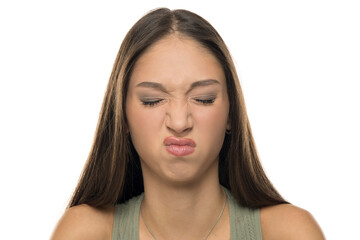 Studio shot of young woman with long brown hair wearing green tank top, grimacing with eyes closed and wrinkled nose, on white background.