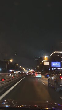 Vertical dashcam view of a car driving along a city highway at night. Urban traffic lights reflecting on the wet asphalt, modern city atmosphere, transportation and travel concept.