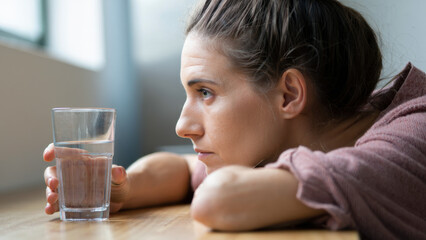 Exhausted woman at home, staring at glass of water, physical fatigue, emotional burnout, mental exhaustion, domestic stillness, silent moment, apathy, sleep deprivation