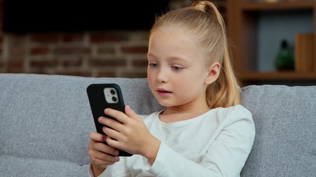 Caucasian little girl kid child daughter looking in mobile phone on sofa at home using smartphone play internet games smiling happy emotional chatting scrolling social networks watching funny video
