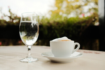White cup of cappuccino with foam and glass of water on an outdoor cafe table, calm atmosphere with natural light and greenery.