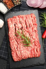 Raw minced meat with rosemary, spices, onion and chili pepper on black textured table, flat lay