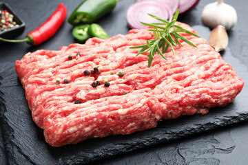 Raw minced meat with rosemary, spices, onion and chili peppers on black textured table, closeup