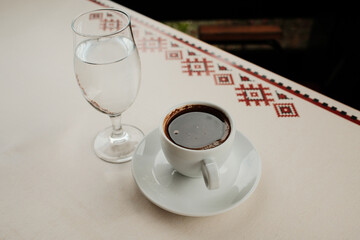 White porcelain cup with strong Turkish black coffee or americano and glass of water served on an outdoor cafe table in natural light.