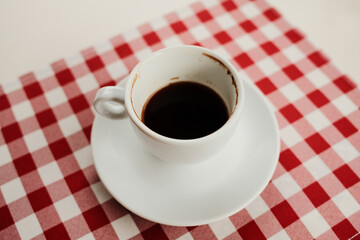 Top view of a white cup filled with black coffee placed on a red and white checkered tablecloth, minimalist breakfast concept.