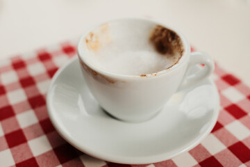 Close-up of a half-finished cappuccino with milk foam in a white cup on a red and white checkered tablecloth, cozy cafe detail.