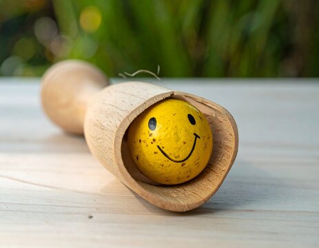 A yellow sphere with a smiling face peeks from a wooden holder on a light wood surface, greenery in the blurred background
