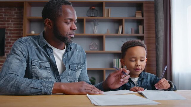 African American family father teaching little son kid boy helping child with writing homework assistance preparing test distant studying education from home learning talking together childcare study