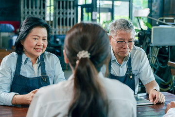 Obraz premium Elderly asian couple restaurant owners sitting with young staff discussing business plan using tablet inside cozy cafe small business teamwork communication collaboration management lifestyle concept