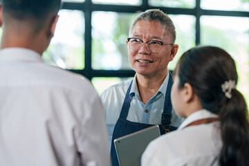 Senior asian cafe owner in denim apron smiling while giving advice to staff during restaurant meeting, teamwork leadership and communication for small business success and customer service improvement