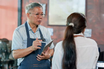 Obraz premium Elderly asian cafe owner in apron holding tablet explaining work process to employee during meeting inside restaurant, small business teamwork communication showing leadership management discussion