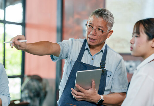 Elderly asian male cafe owner in apron holding tablet while instructing employee during restaurant meeting, teamwork leadership in small business collaboration for staff training and service