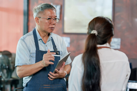 Elderly asian cafe owner in apron holding tablet explaining work process to employee during meeting inside restaurant, small business teamwork communication showing leadership management discussion