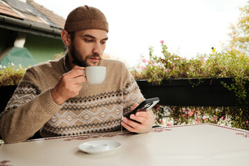 Young man in brown sweater holding coffee cup and using smartphone at outdoor cafe table, casual modern lifestyle scene