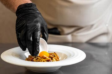 Chef plating gourmet pasta with raw egg yolk in professional kitchen. Culinary presentation, close-up of hands, black glove, stainless steel surface.