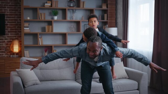 Happy African American family playing piggyback at home joyful carefree father dad riding on back little son spinning play with two kids boys sons children having fun fooling around jumping on couch