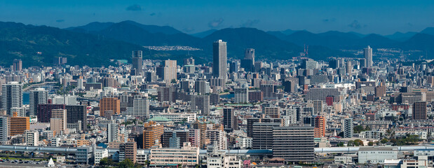 Obraz premium Aerial view shows Hiroshima, Japan, with mid and high rises, a broad rail corridor, and dark blue mountains. Clear daylight, cool tones, urban density against ridgelines.