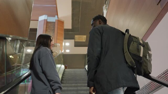 Two business colleagues riding up escalator inside modern building, engaging in cheerful conversation, wearing casual jackets and carrying shopping bags and backpack