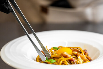 Chef garnishing gourmet spaghetti carbonara with egg yolk, bacon, cheese, and basil in a white bowl. Close-up of plating process with black glove and metal tongs.