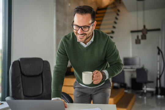 Smiling business man working remotely using laptop and drinking coffee - Powered by Adobe