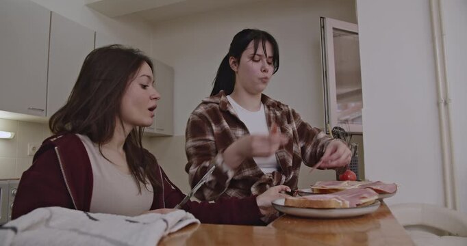 Two female roommates enjoy making and eating ham and tomato sandwiches in their kitchen. They are happy and having fun.