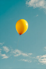 Fototapeta premium Yellow balloon is soaring high in a clear blue sky, surrounded by soft white clouds, creating a cheerful and uplifting atmosphere