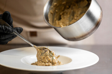 Chef plating creamy risotto with herbs from stainless pot into white bowl in professional kitchen. Culinary preparation, gourmet food, restaurant service concept.