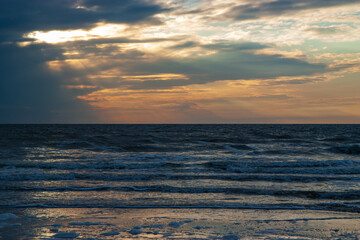 Dramatic sunset over the North Sea near Texel, with sun rays bursting through dark, colorful clouds and gentle waves reaching the sandy shore