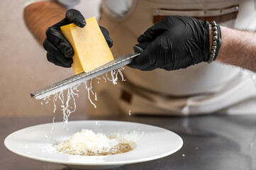 Chef grating cheese over plated pasta in professional kitchen. Close-up of hands in gloves, apron, and bracelet. Culinary preparation with fresh ingredients.