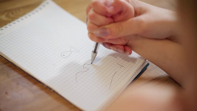 Child hand holds pen while practicing drawing shapes on squared notebook page during learning activity at home, showing early writing skills, concentration, and creativity with closeup view