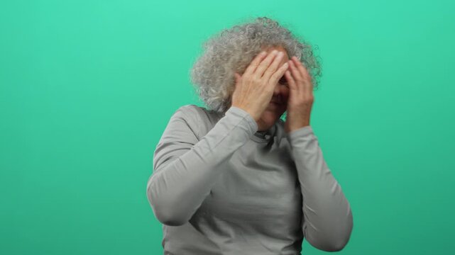 Woman expressing fear with hands raised against a green background, featuring an elderly woman with grey hair in an isolated indoor setting.