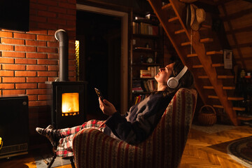 A woman wearing headphones holds a smartphone and relaxes while sitting in a chair next to a stove in the evening