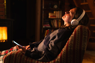 A woman wearing headphones holds a smartphone and relaxes while sitting in a chair next to a stove in the evening