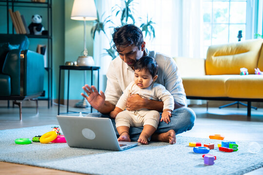 Indian Father and infant boy share laptop video call while sitting on carpet in modern home - Powered by Adobe