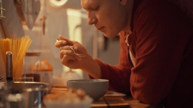 A person is savoring pure pumpkin from a bowl while sitting at a kitchen counter. The warm lighting adds a cozy atmosphere to the evening meal in a home kitchen