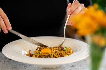 Close-up of spaghetti carbonara with raw egg yolk, bacon, and herbs. A person mixes the pasta with fork and knife. Warm lighting, gourmet presentation, casual dining atmosphere.