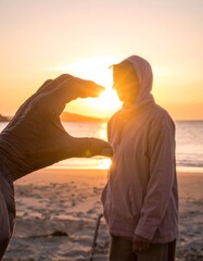 A hand frames the sun near a hooded figure on a beach