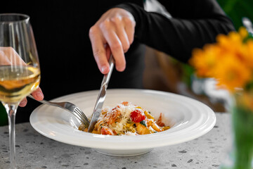 Person eating pasta with fork and knife at a cozy table setting, garnished with cheese and cherry tomatoes, accompanied by white wine and yellow flowers. Warm dining atmosphere.