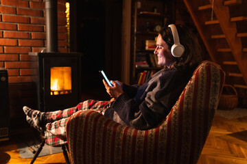A woman wearing headphones holds a smartphone while sitting in a chair next to a stove in the evening