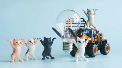Cute toy kittens act as electricians while a toy loader lifts a big LED bulb; playful scene about energy saving, electrification and STEM learning on blue. Photo