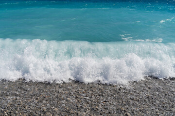 Close-up of a beautiful turquoise sea wave with white foam crashing on a pebble beach in Nice, France.