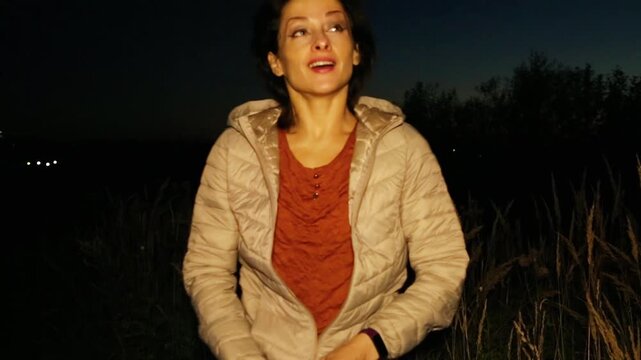 Expression on the face of a frozen brunette walking across a field against the backdrop of a bright autumn sunset and a city in cold weather, she zips up her jacket and pulls the hood over her head
