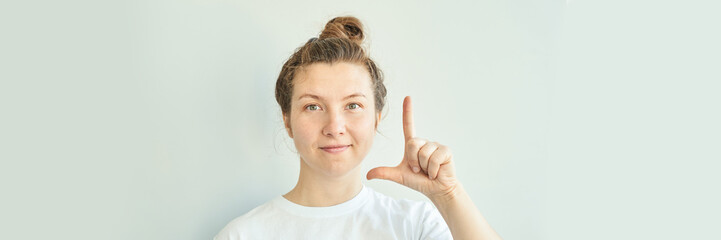 Caucasian young female with a topknot making l sign gesture in a white shirt against light background.